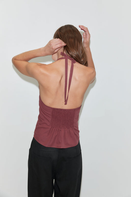 A woman with long brown hair faces away as she ties the strap of a Folded Top - Rust Stripe by Deiji Studios. She wears loose black pants and stands against a plain white background.