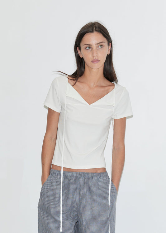 A cropped view of a woman wearing the Linen Ease Trouser - Indigo Check by Deiji Studios, indigo micro-check wide-leg linen trousers with a drawcord waist, and a white tie-back tee against a plain white background.