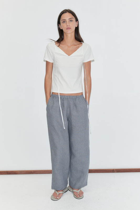 A woman with long dark hair stands against a plain white background with her hands in her pockets, wearing the Linen Ease Trouser - Indigo Check by Deiji Studios, indigo micro-check wide-leg linen trousers, with a white tie-back tee and grey flip-flops.