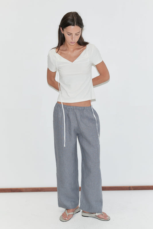 A woman with long dark hair stands against a plain white background looking down, wearing the Linen Ease Trouser - Indigo Check by Deiji Studios, indigo micro-check wide-leg linen trousers, with a white tie-back tee and flip-flops.