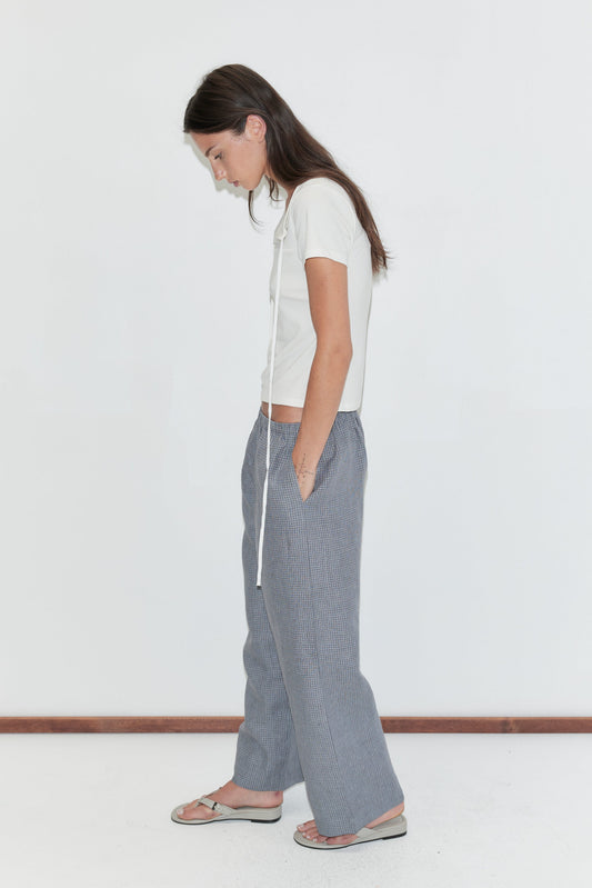 A woman with long dark hair stands in profile against a plain white background wearing the Linen Ease Trouser - Indigo Check by Deiji Studios, indigo micro-check wide-leg linen trousers, with a white tie-back tee and flip-flops.