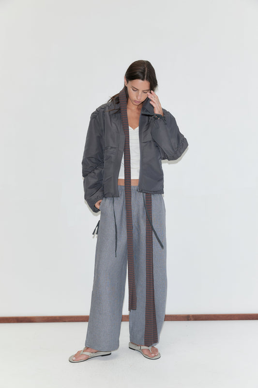 A woman with long dark hair stands against a plain white background wearing the Linen Ease Trouser - Indigo Check by Deiji Studios, indigo micro-check wide-leg linen trousers, with a white top and a slate grey gathered jacket.