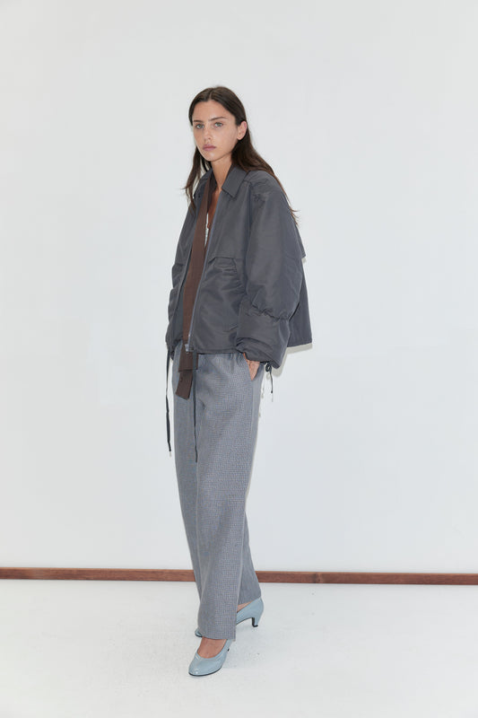 A woman with long dark hair stands against a plain white background wearing the Linen Ease Trouser - Indigo Check by Deiji Studios, indigo micro-check wide-leg linen trousers, with a slate grey gathered jacket and pale blue heels.