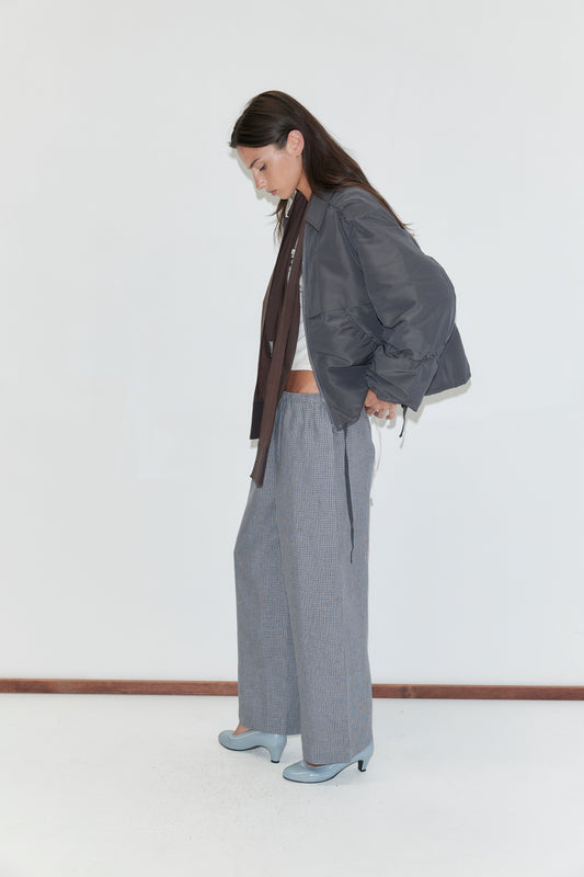 A woman with long dark hair stands in profile against a plain white background wearing the Linen Ease Trouser - Indigo Check by Deiji Studios, indigo micro-check wide-leg linen trousers, with a slate grey gathered jacket and pale blue heels.