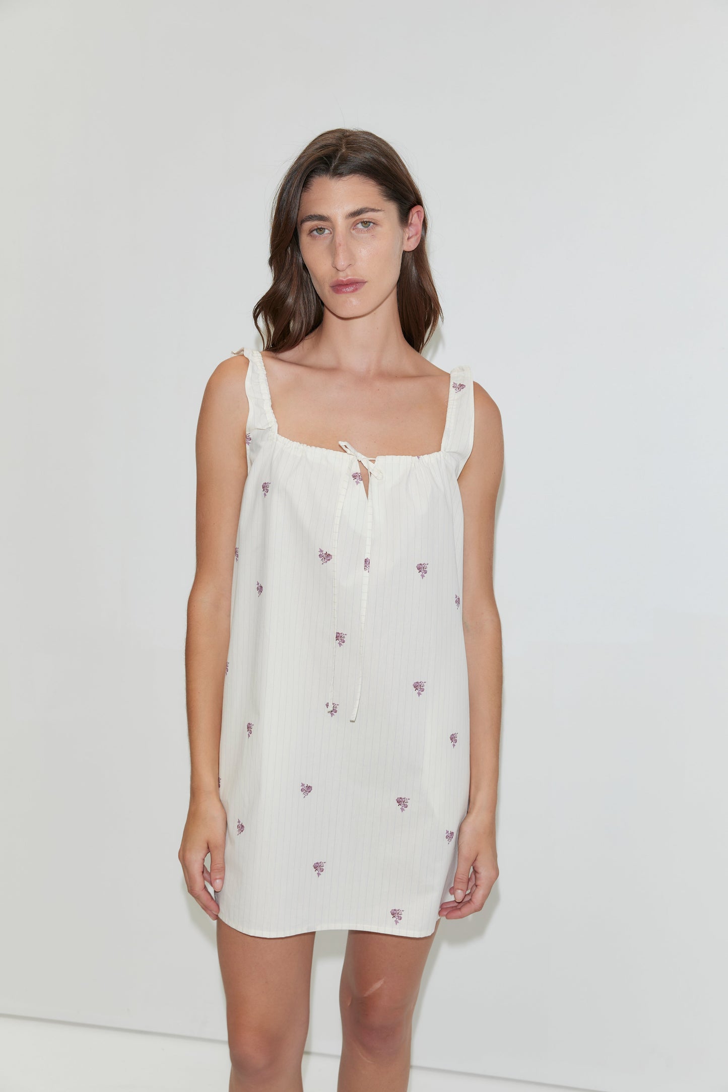 A woman stands against a plain white background wearing the Bow Dress - Floral Stripe, a white mini-length dress featuring small purple floral prints and ruffled shoulder straps.