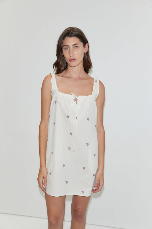 A woman stands against a plain white background wearing the Bow Dress - Floral Stripe, a white mini-length dress featuring small purple floral prints and ruffled shoulder straps.