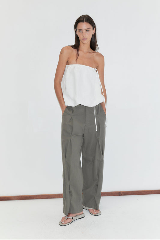 A woman with long dark hair stands against a plain white background wearing the Bubble Top - White by Deiji Studios, a strapless white linen top with a gathered bubble hem, with olive check wide-leg trousers and grey flip-flops.