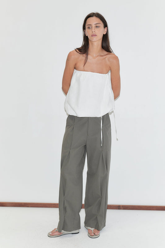 A woman with long dark hair stands against a plain white background wearing the Bubble Top - White by Deiji Studios, a strapless white linen top with a gathered bubble hem, with olive check wide-leg trousers and grey flip-flops.