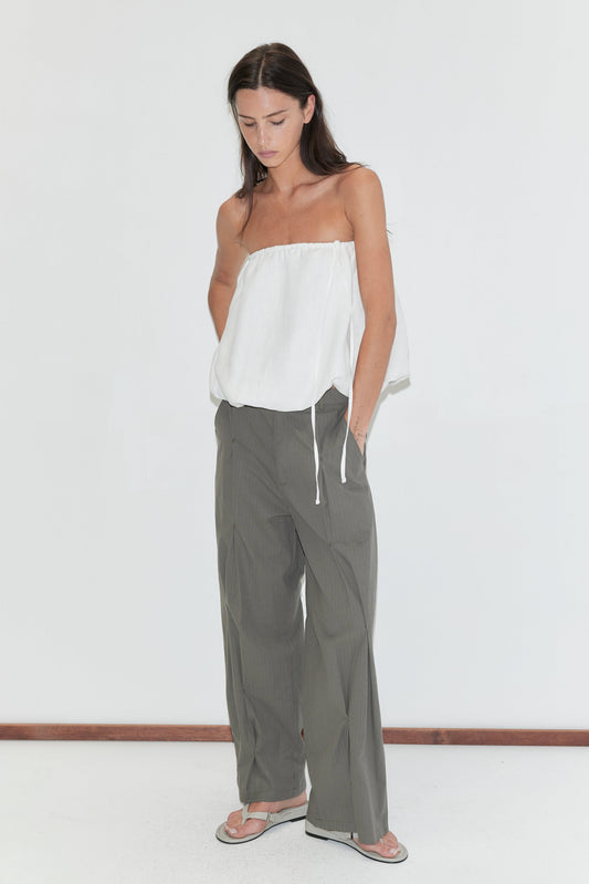 A woman with long dark hair stands against a plain white background wearing the Multi Pleat Pant - Moss Stripe by Deiji Studios, moss-green fine-check wide-leg pleated trousers, with a white strapless top and flip-flops.