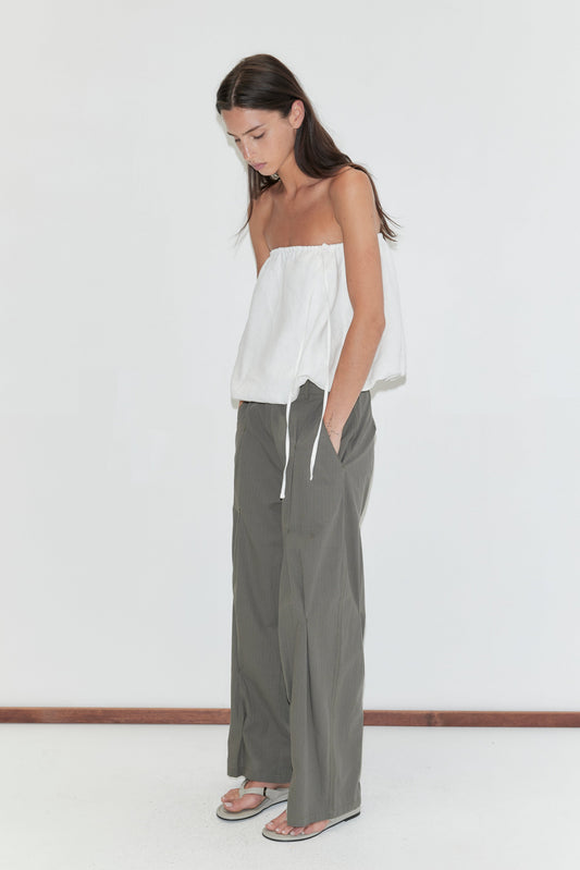 A woman with long dark hair stands in profile against a plain white background wearing the Multi Pleat Pant - Moss Stripe by Deiji Studios, moss-green fine-check wide-leg pleated trousers, with a white strapless top and flip-flops.