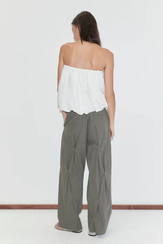 A woman with long dark hair stands with her back to the camera against a plain white background wearing the Bubble Top - White by Deiji Studios, a strapless white linen top with a gathered bubble hem, with olive check wide-leg trousers.