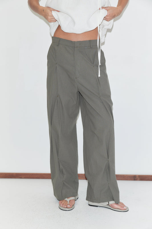 A cropped view of the Multi Pleat Pant - Moss Stripe by Deiji Studios, moss-green fine-check wide-leg pleated trousers with a waistband, worn with a white top and flip-flops against a plain white background.