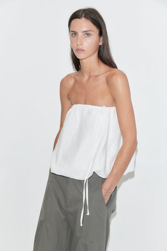 A woman with long dark hair stands against a plain white background wearing the Bubble Top - White by Deiji Studios, a strapless white linen top with a gathered edge and drawcord side tie, paired with olive check wide-leg trousers.