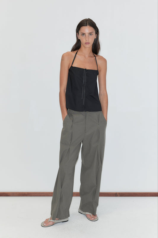 A woman with long dark hair stands against a plain white background wearing the Simple Tie Top - Black by Deiji Studios, a black halter top with a gathered neckline and front tie, with grey check pleated trousers and flip-flops.