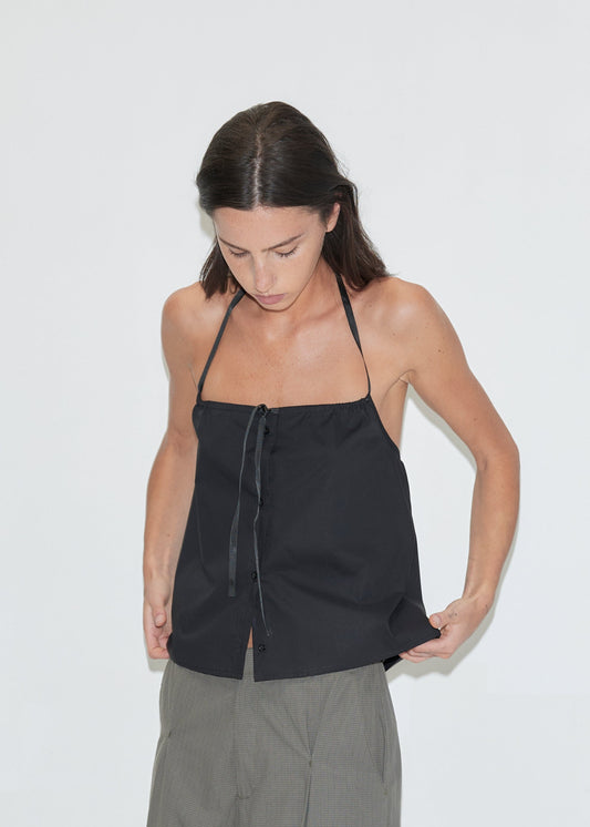 A cropped view of a woman looking down, wearing the Simple Tie Top - Black by Deiji Studios, a black halter top with a gathered neckline and drawstring front tie, with grey check trousers against a plain white background.