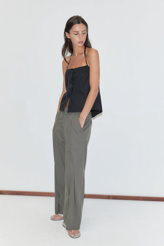 A woman with long dark hair stands against a plain white background wearing the Simple Tie Top - Black by Deiji Studios, a black halter top with a front tie, with grey check pleated trousers and flip-flops.