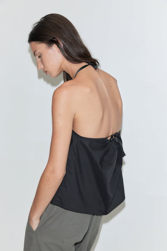 A woman with long dark hair stands in profile against a plain white background wearing the Simple Tie Top - Black by Deiji Studios, a black halter top with an open back and tie fastening, with grey check trousers.