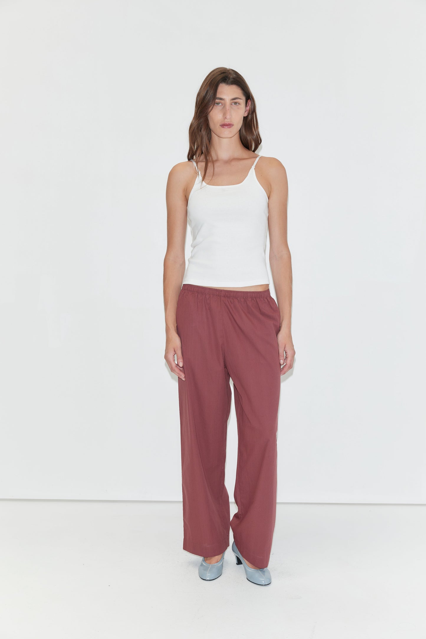 A woman with long brown hair and a neutral expression stands against a plain white background, wearing a white tank top and the Ease Trouser - Rust Stripe by Deiji Studios. She pairs the outfit with light blue shoes.