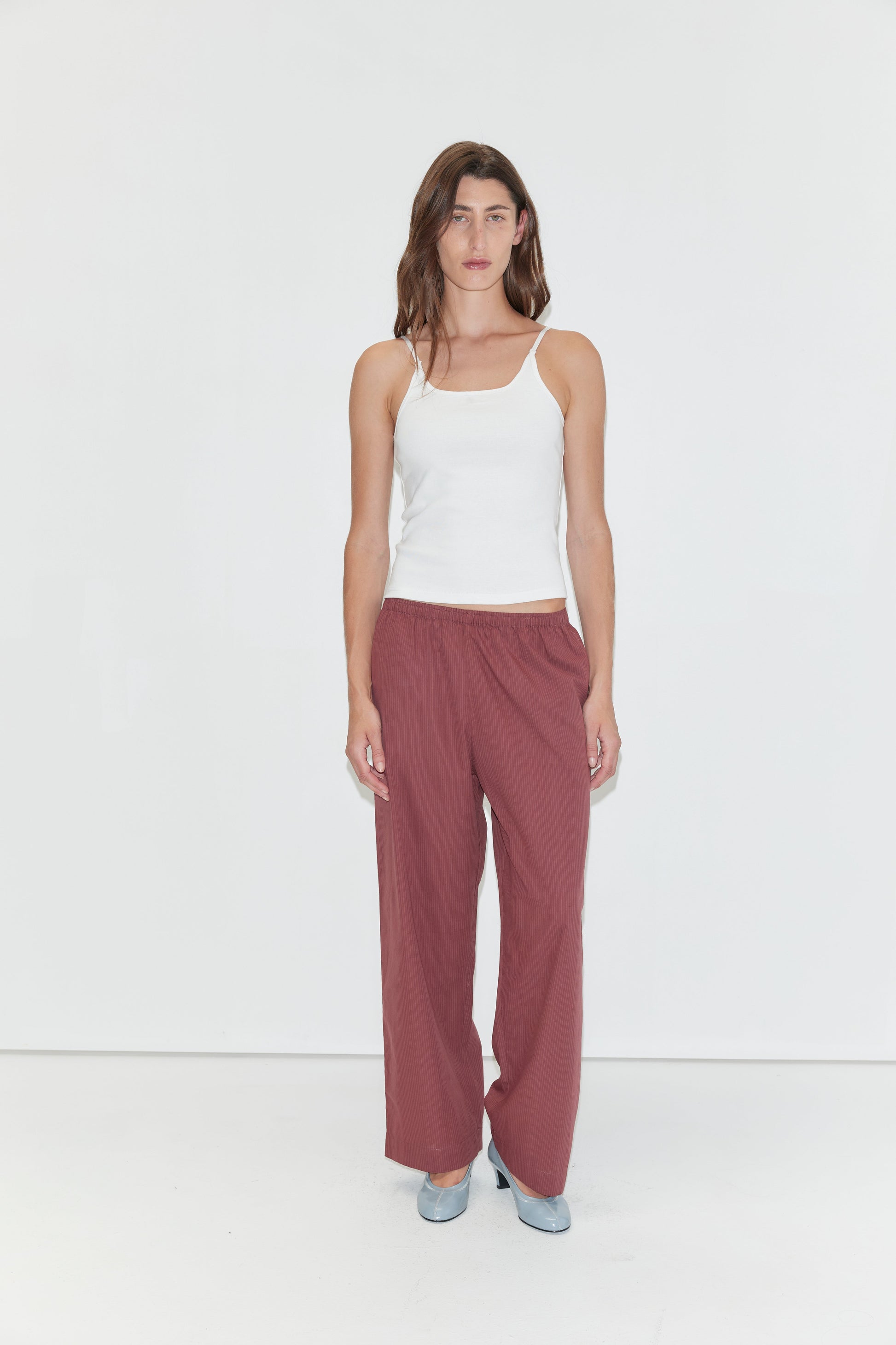 A woman with long brown hair and a neutral expression stands against a plain white background, wearing a white tank top and the Ease Trouser - Rust Stripe by Deiji Studios. She pairs the outfit with light blue shoes.