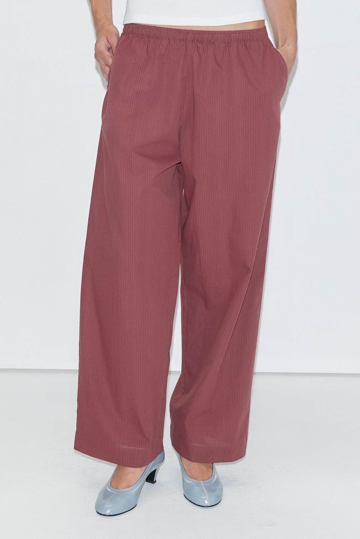 A person wearing the Ease Trouser - Rust Stripe from Deiji Studios with hands in pockets, paired with light blue heeled shoes against a plain white background.