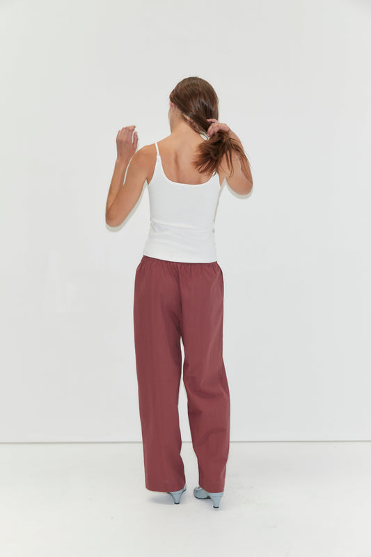 A woman stands facing a white wall, holding her brown hair in a ponytail. She wears the Rib Jersey Tank in White from Deiji Studios, loose maroon pants, and light blue heels.