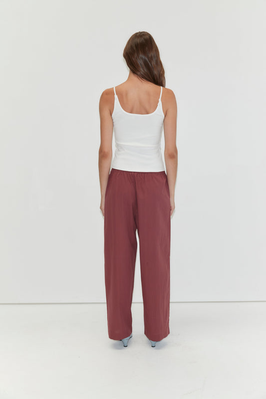 A woman with long brown hair, wearing a white tank top and Deiji Studios Ease Trouser in Rust Stripe, stands facing away from the camera against a plain white background.