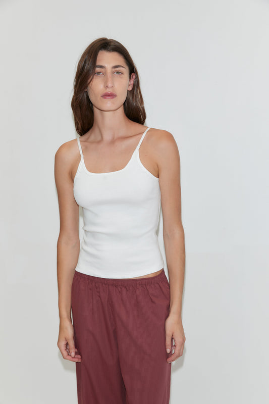 A woman with long brown hair wears the Rib Jersey Tank in White by Deiji Studios and maroon pants as she stands against a plain light-colored background.