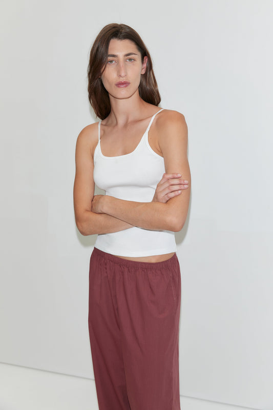 A person with long brown hair wearing a white tank top and the Ease Trouser - Rust Stripe stands against a plain white background with arms crossed, looking at the camera.