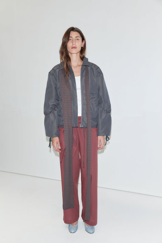 A person stands against a white background wearing a gray bomber jacket, a white top, the Ease Trouser - Rust Stripe from Deiji Studios, a long striped scarf, and light blue shoes. They have long brown hair and a neutral expression.