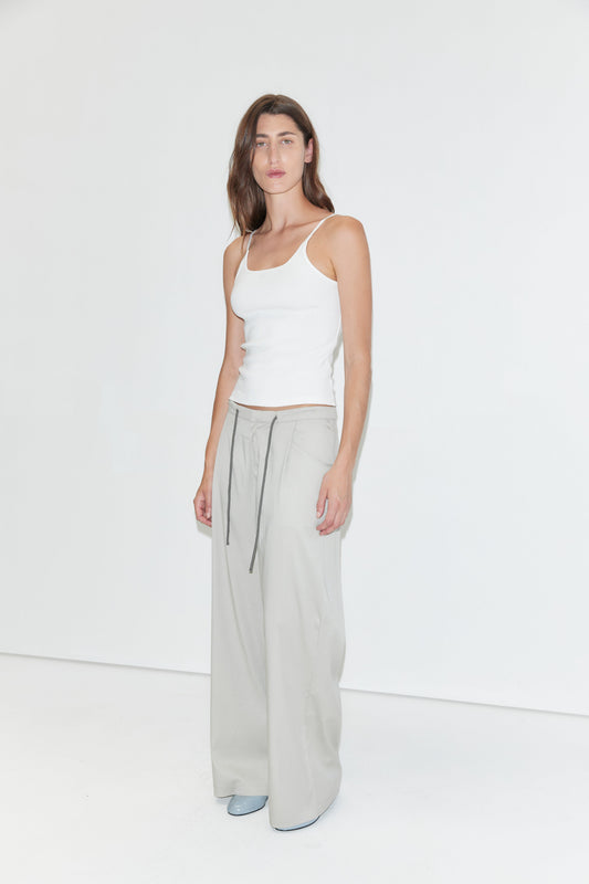 A person stands in a minimalist white space, wearing the Rib Jersey Tank in white from Deiji Studios and loose, light gray pants with ties, paired with gray shoes. They have long brown hair and a neutral expression.