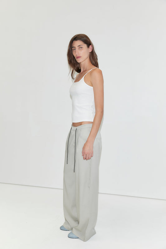 A woman with long brown hair wears a white tank top, the Tencel Pant - Grey from Deiji Studios—loose light gray wide-leg pants with drawstrings—and blue shoes, standing against a plain white background.