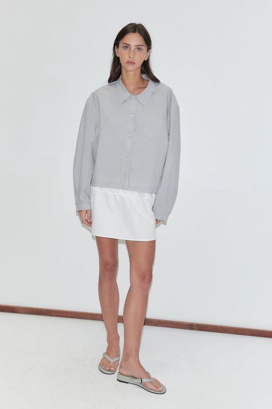 A woman with long dark hair stands against a plain white background wearing the Flounce Layer Mini Skirt - White by Deiji Studios, a white low-rise mini skirt with a flounce waist, with a pale grey striped boxy shirt and flip-flops.