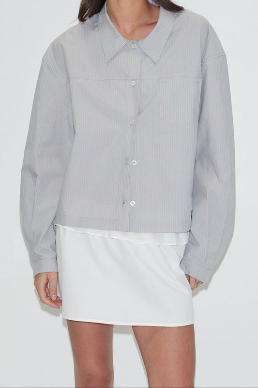 A cropped front view of the Boxy Shirt - Cloud Stripe by Deiji Studios, a pale grey-blue fine-striped boxy shirt with a collar, front buttons and chest pocket, worn over a white mini skirt against a plain white background.