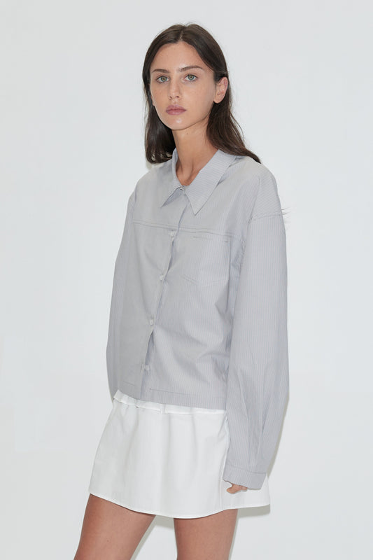 A woman with long dark hair stands against a plain white background wearing the Boxy Shirt - Cloud Stripe by Deiji Studios, a pale grey-blue fine-striped boxy shirt, over a white flounce mini skirt with grey flip-flops.