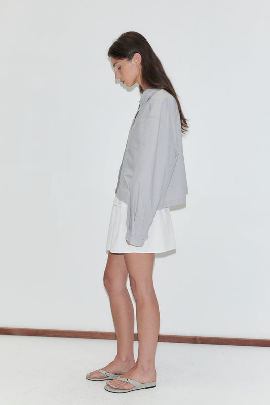 A woman with long dark hair stands in profile against a plain white background wearing the Flounce Layer Mini Skirt - White by Deiji Studios, a white low-rise mini skirt with a flounce waist, with a pale grey striped boxy shirt and flip-flops.