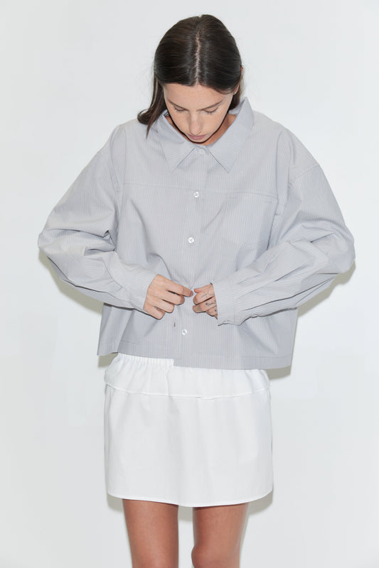 A cropped view of a woman looking down, wearing the Flounce Layer Mini Skirt - White by Deiji Studios, a white low-rise mini skirt with a flounce layer at the waist, and a pale grey striped boxy shirt against a plain white background.