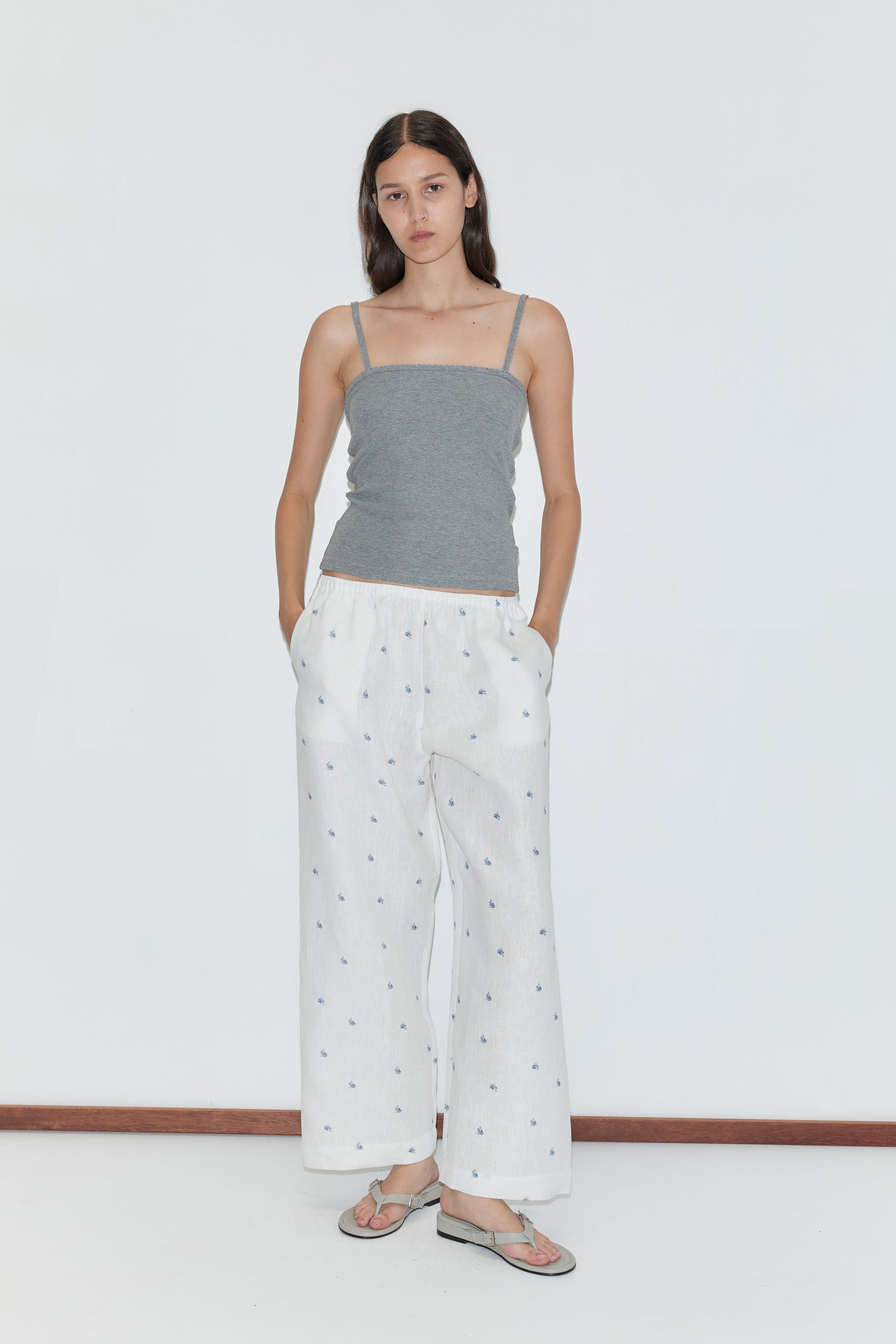 A woman with straight brown hair stands against a plain white background, wearing the Linen Ease Trouser—loose white pants with a subtle print—paired with a gray spaghetti strap top and sandals, her hands in her pockets.