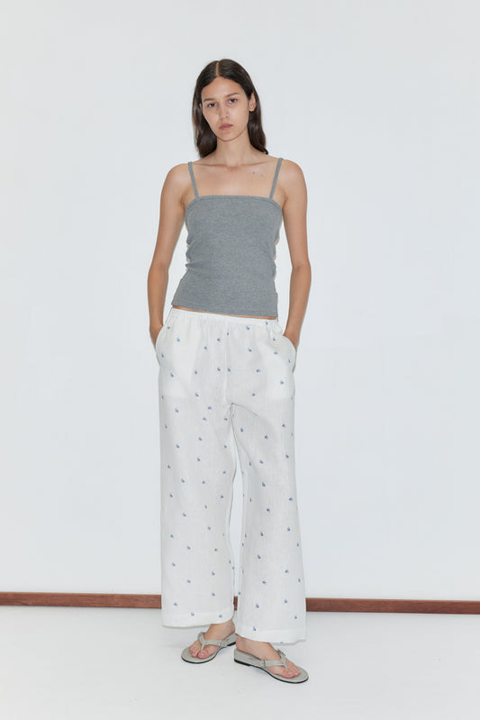 A woman with straight brown hair stands against a plain white background, wearing the Linen Ease Trouser—loose white pants with a subtle print—paired with a gray spaghetti strap top and sandals, her hands in her pockets.