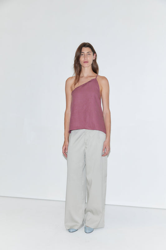A person stands against a plain white background wearing the Half Halter - Brick linen one-shoulder top from Deiji Studios, paired with loose light gray pants and light blue shoes.