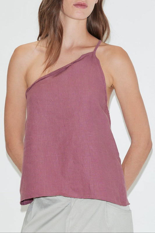 A woman wearing the Half Halter - Brick, a sleeveless linen one-shoulder top from Deiji Studios, paired with light-colored pants and standing against a plain white background.