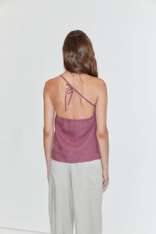 A woman stands facing away, wearing the Half Halter top in Brick by Deiji Studios and light gray pants. Her long brown hair is down, and the background is plain white.