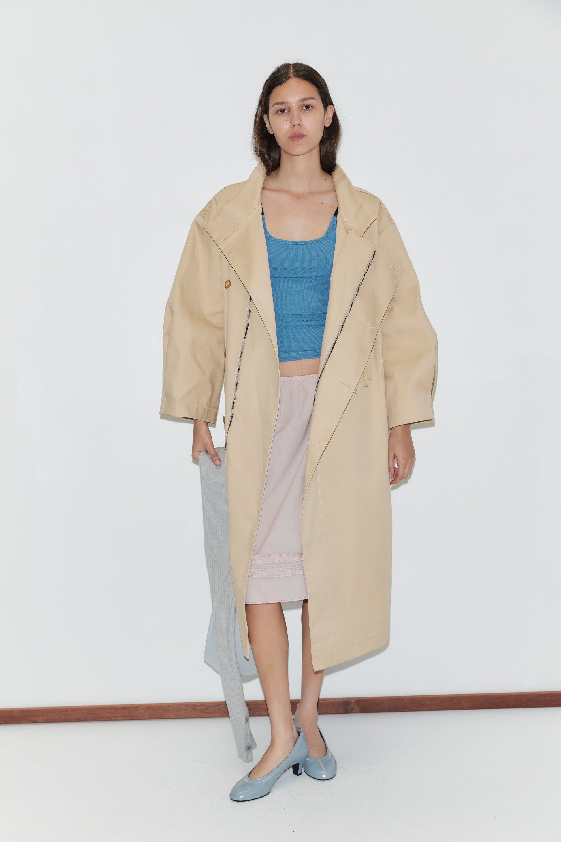 A woman stands against a white wall, wearing a loose beige trench coat over the Rib Jersey Tank in Azure by Deiji Studios and a pale pink skirt. She holds a gray garment and wears light blue shoes.