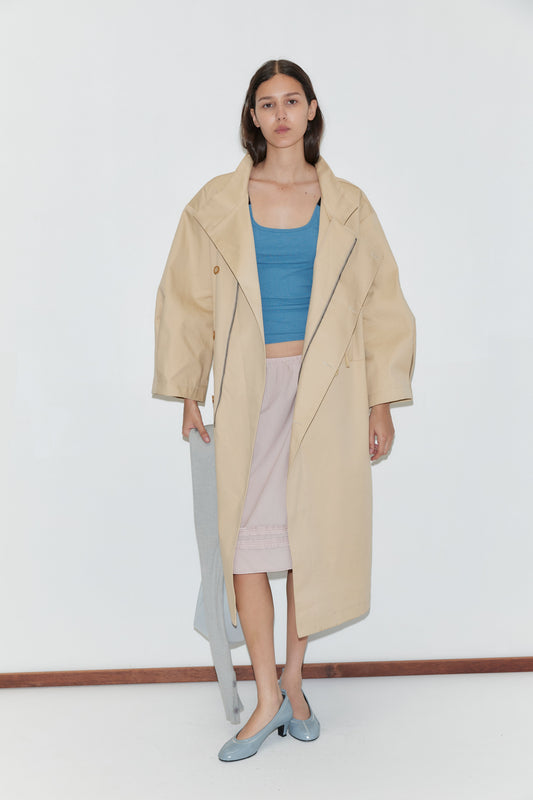 A woman stands against a white wall, wearing a loose beige trench coat over the Rib Jersey Tank in Azure by Deiji Studios and a pale pink skirt. She holds a gray garment and wears light blue shoes.