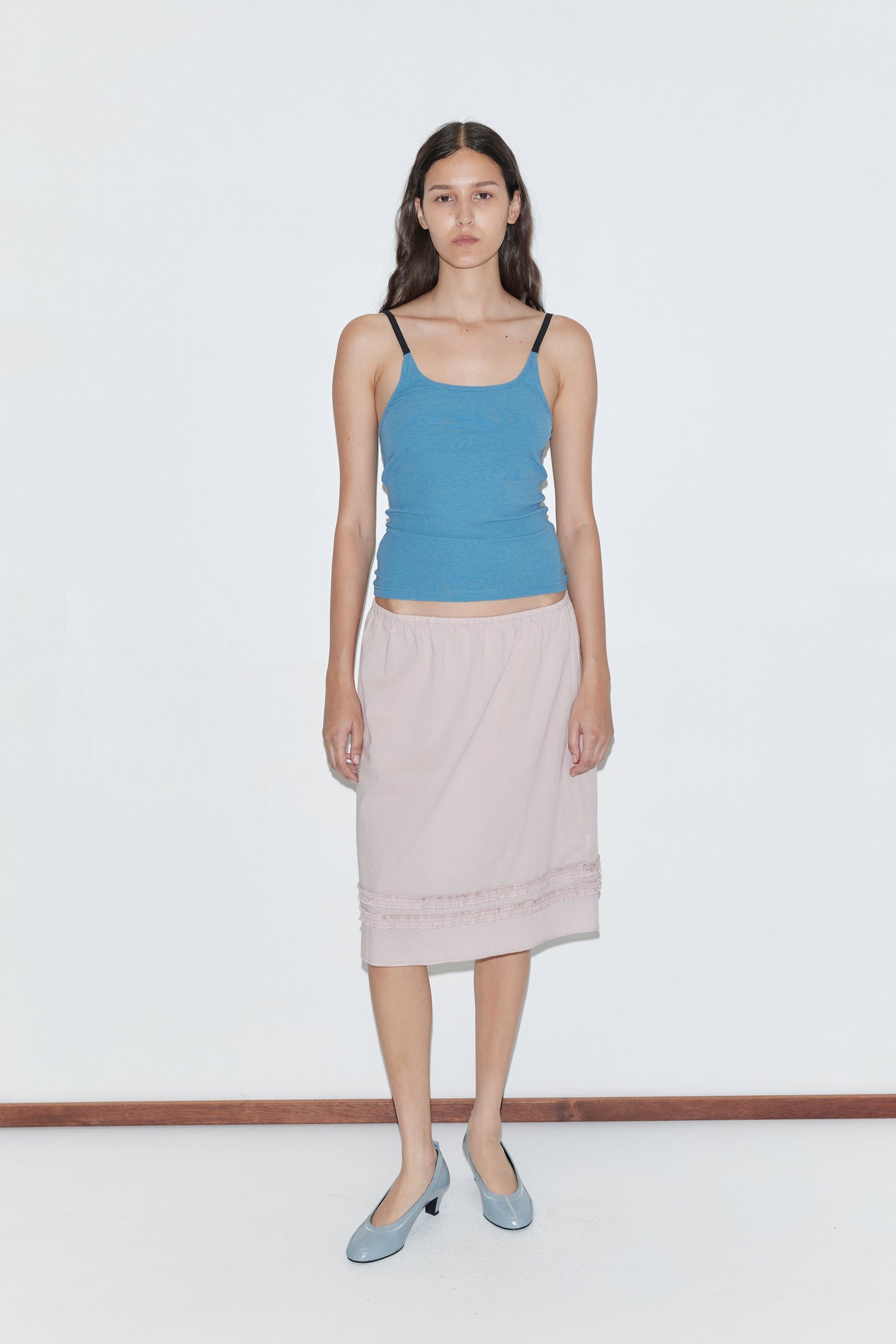 A woman with long dark hair stands against a plain white background, wearing the Rib Jersey Tank - Azure by deiji studios, a light pink knee-length skirt, and light blue shoes. She looks straight ahead with a neutral expression.