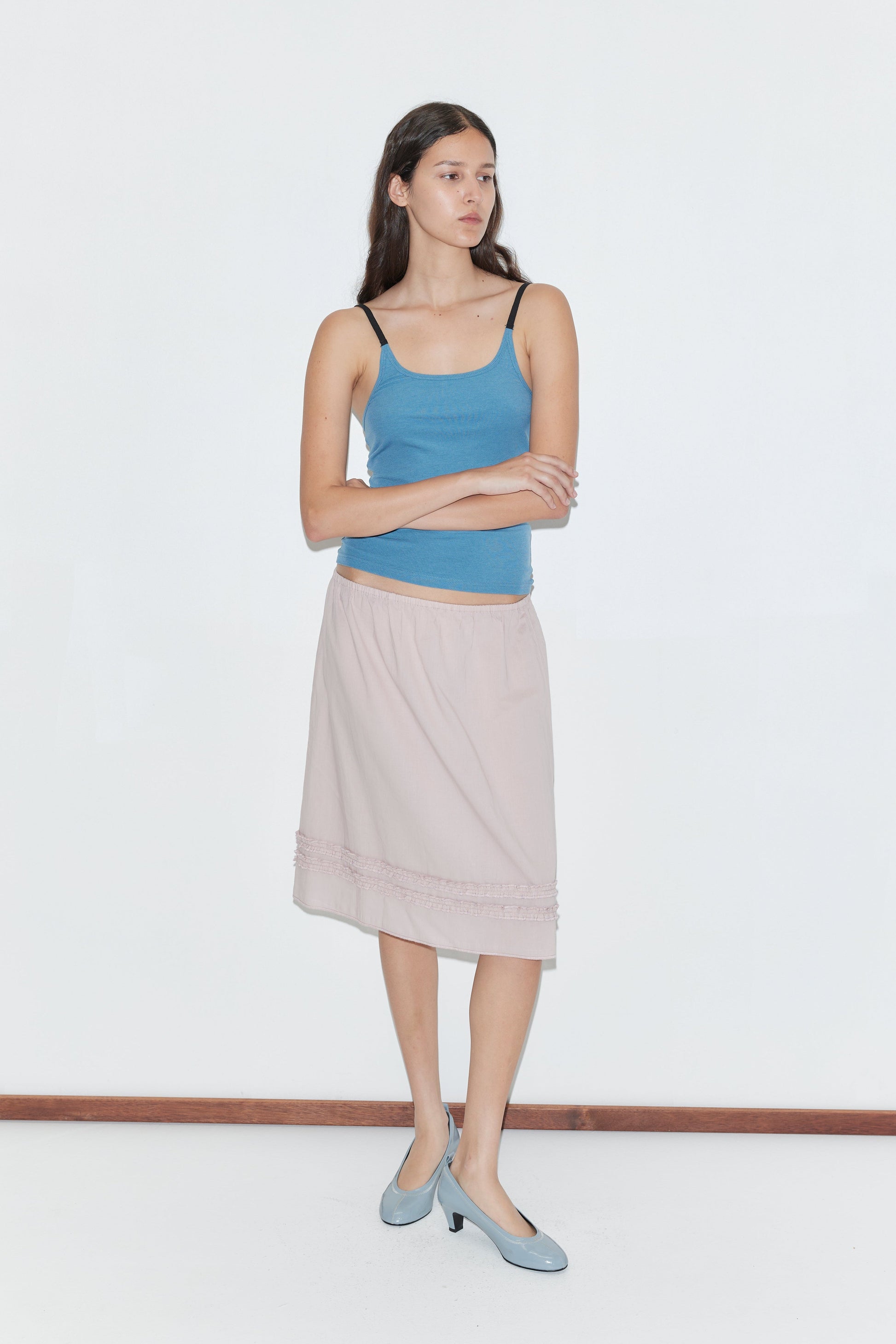 A woman with long brown hair stands against a plain white background, wearing the Rib Jersey Tank - Azure by Deiji Studios, a light pink knee-length skirt, and light blue shoes. She crosses her arms and looks to the side.