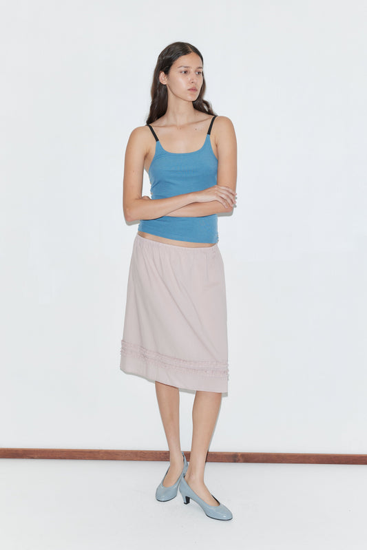 A woman with long brown hair stands against a plain white background, wearing the Rib Jersey Tank - Azure by Deiji Studios, a light pink knee-length skirt, and light blue shoes. She crosses her arms and looks to the side.