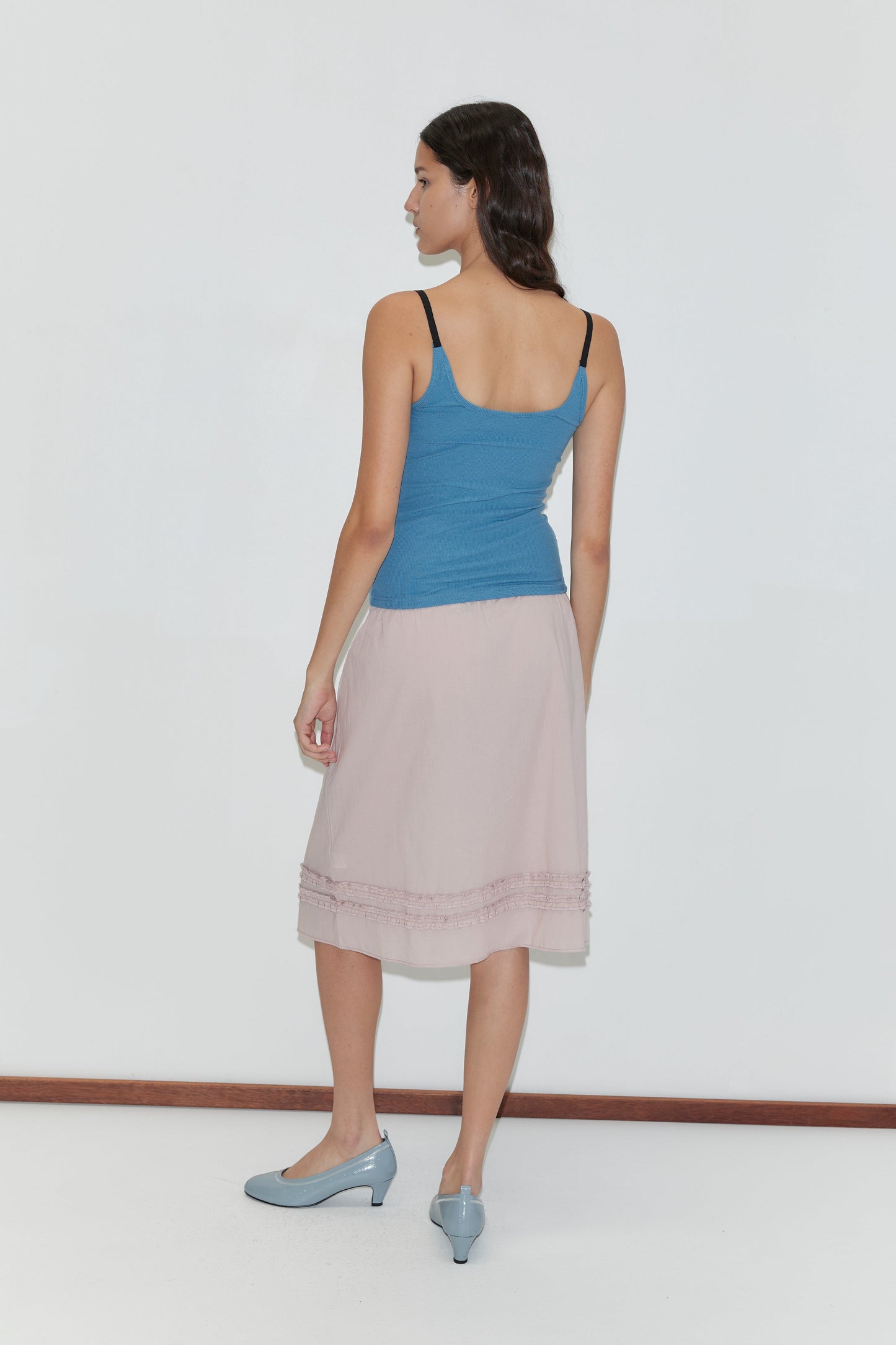 A woman with long dark hair faces away, wearing a Deiji Studios Rib Jersey Tank - Azure with a square neckline, a light pink knee-length skirt, and light blue heels against a plain white background.