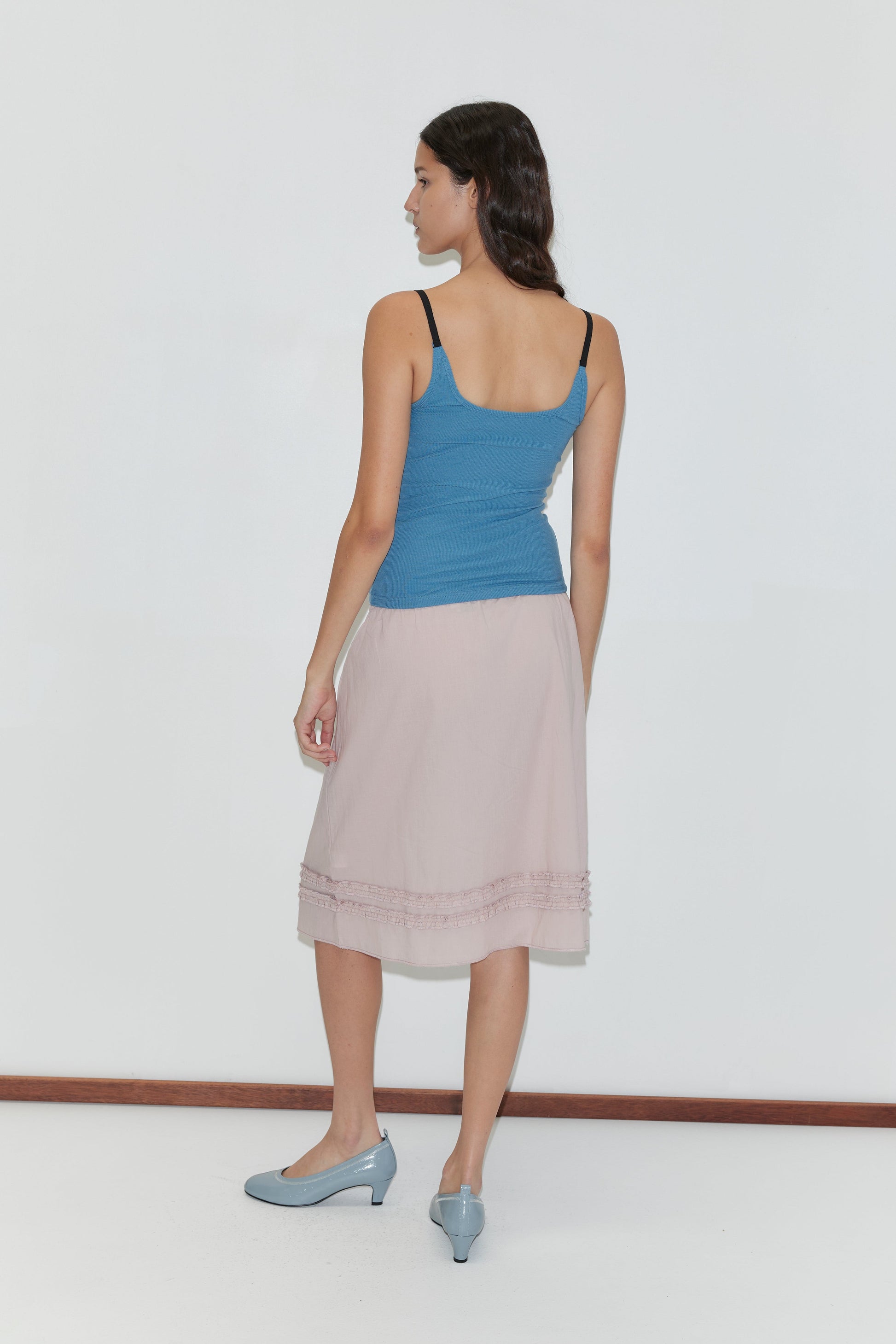 A woman with long dark hair faces away, wearing a Deiji Studios Rib Jersey Tank - Azure with a square neckline, a light pink knee-length skirt, and light blue heels against a plain white background.