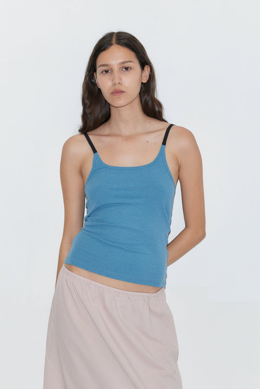 A young woman with long dark hair stands against a white background, wearing the Rib Jersey Tank - Azure by Deiji Studios with black straps and a light beige skirt. She has a neutral expression and relaxed posture.
