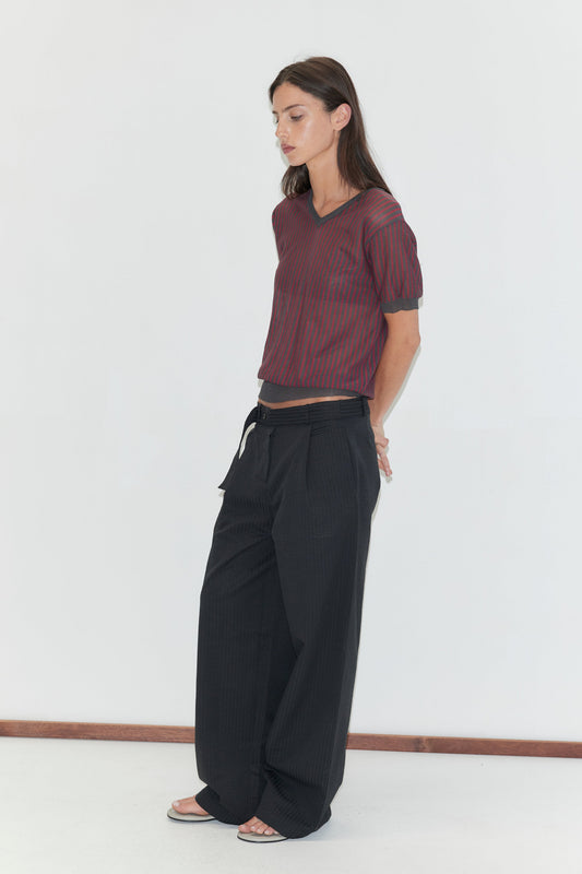 A woman with long dark hair stands in profile against a plain white background wearing the Cross Neck Tee - Red Stripe by Deiji Studios, a fine red and charcoal striped knit tee, with black wide-leg trousers and flip-flops.